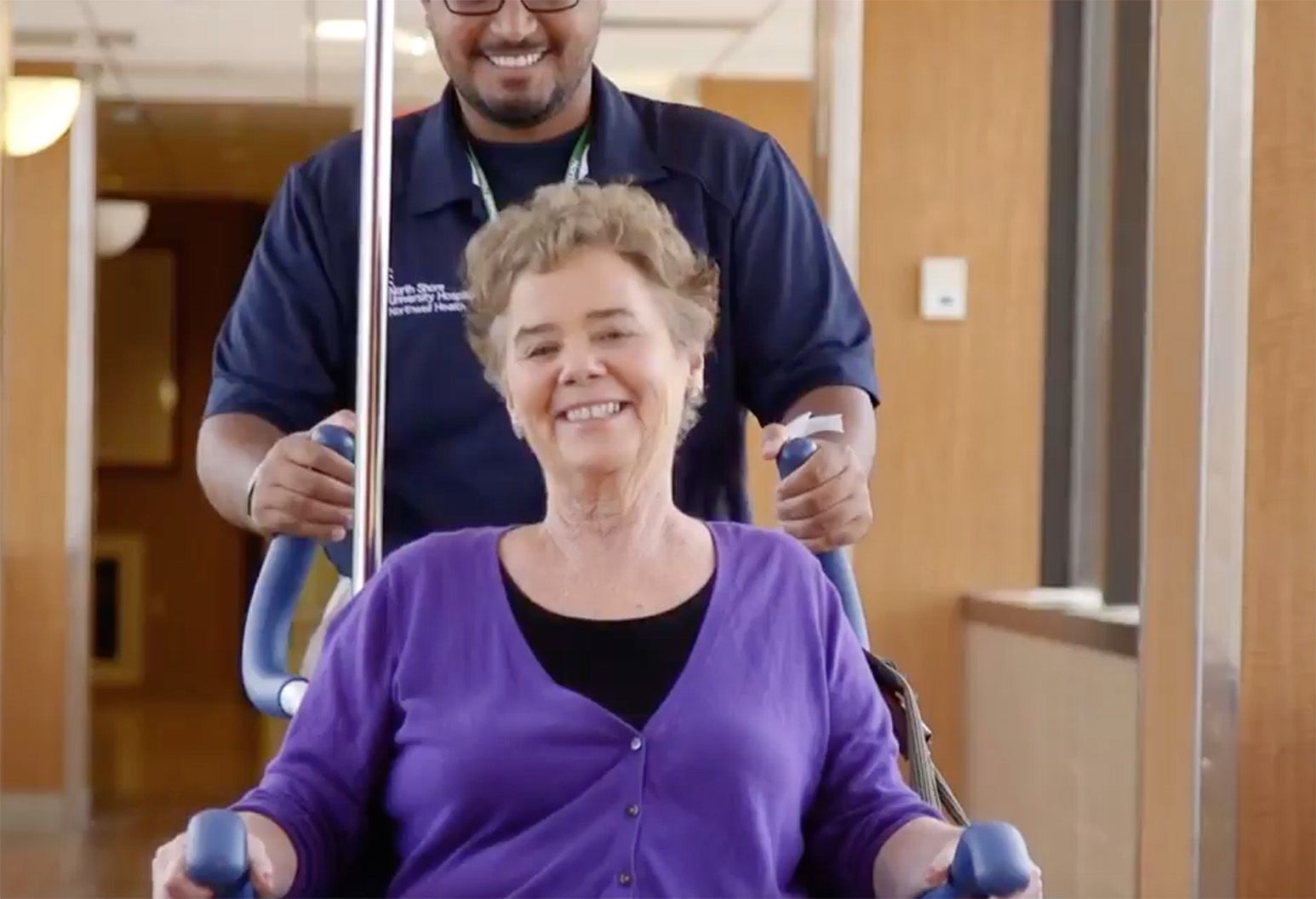 A smiling man in glasses and a navy polo shirt pushes a smiling elderly woman in a wheelchair.