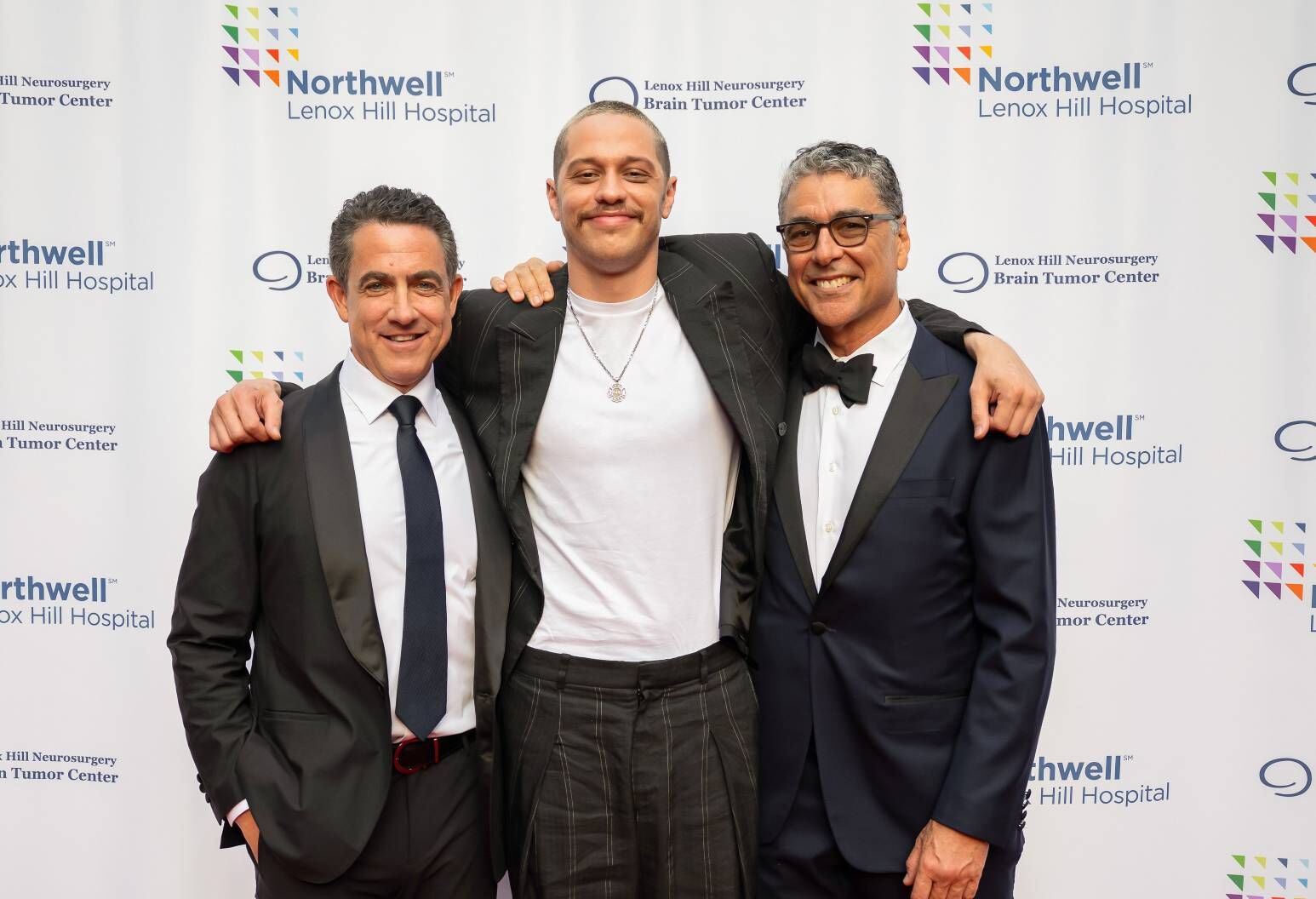 A man wearing a white t-shirt and black jacket stands in between two other men dressed in black tuxedos.